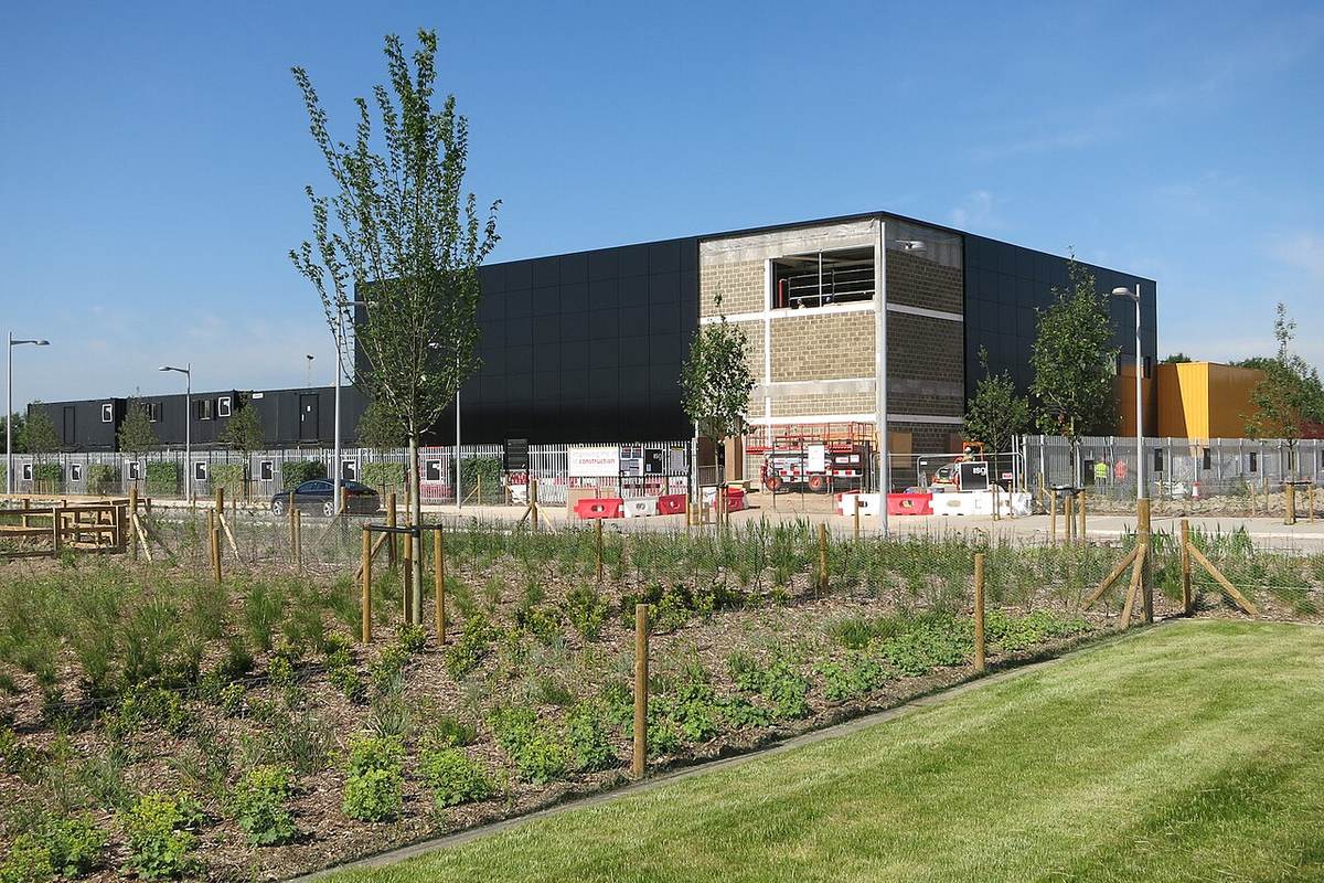 Large dark-clad data centre building under construction behind a fence, with young trees and planting in front
